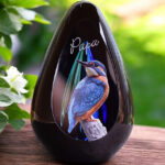 een glazen urn met een blauwe askamer en een afbeelding van een ijsvogeltje en een naam op de urn bewerkt