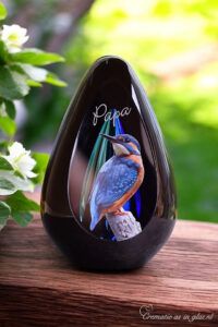 een glazen urn met een blauwe askamer en een afbeelding van een ijsvogeltje en een naam op de urn bewerkt