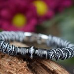 Heren gedenkarmband van zwart-wit paracord met zwarte design bedels waarvan de middelste rvs bedel crematie-as zichtbaar bevat.