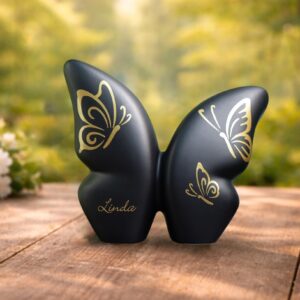 Zwarte keramische vlinder urn op houten tafel in bos, met goudfolie vlinders en ruimte voor naam, geschikt voor crematie-as.